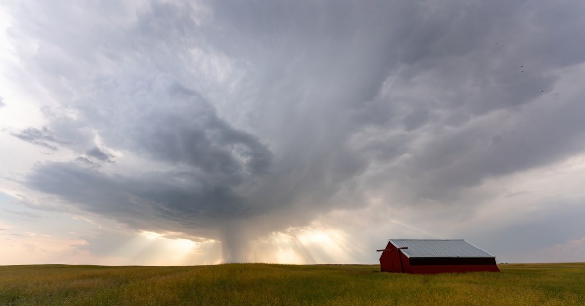 A red barn sits in a field of tall grass. A rainstorm is gathering in the distance with large dark clouds.