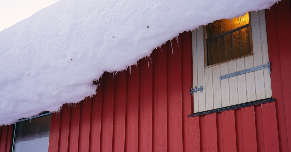 A close-up of a red barn. A large amount of snow is accumulated on the roof and leaning over the side of the structure.