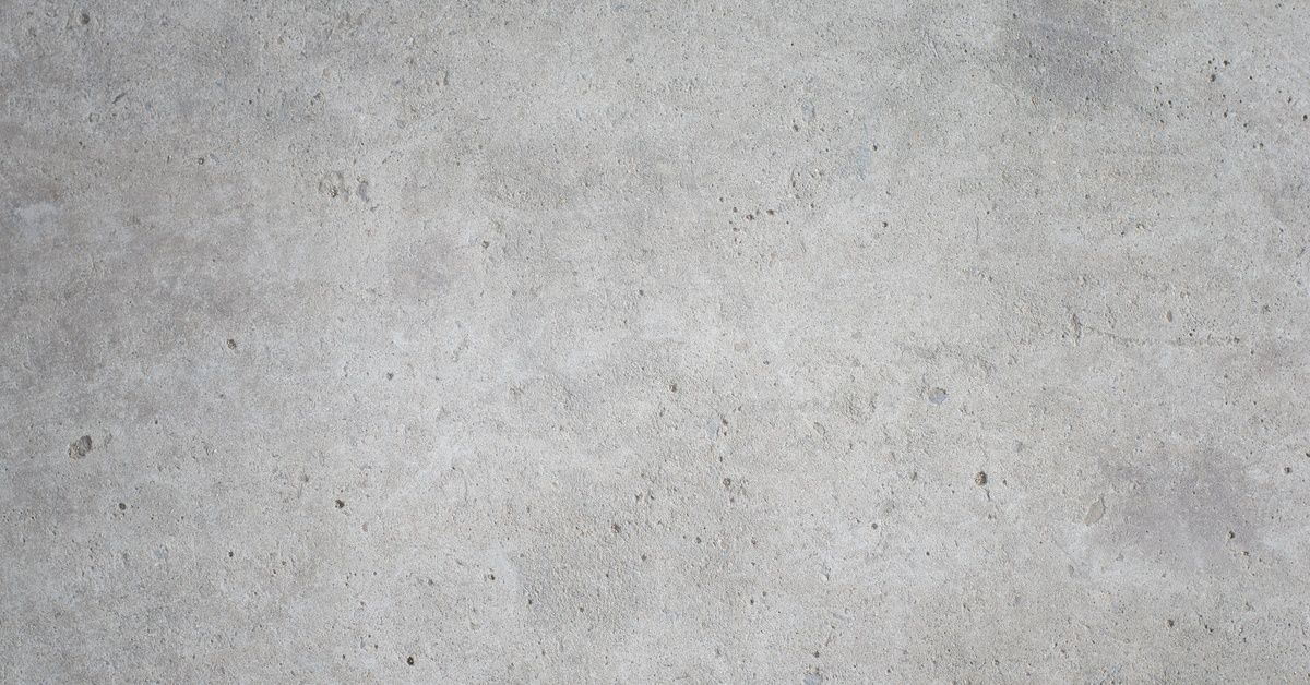 A close-up of a light gray concrete surface with a granular texture, scattered dark specks, and subtle lighter patches.