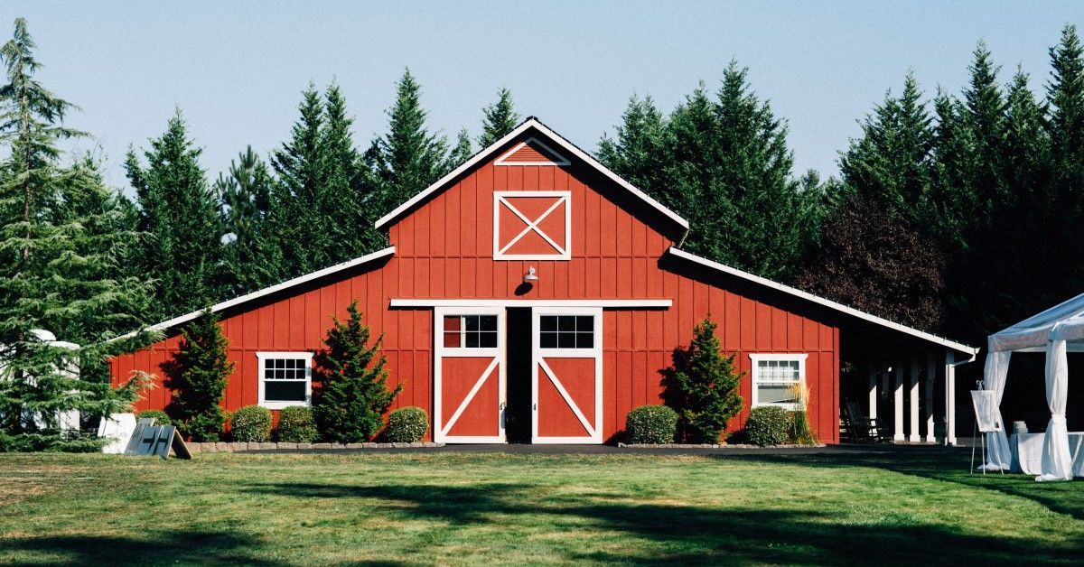 A large, bright red barn with white trim and sliding doors sits in a grassy field with a forest of p