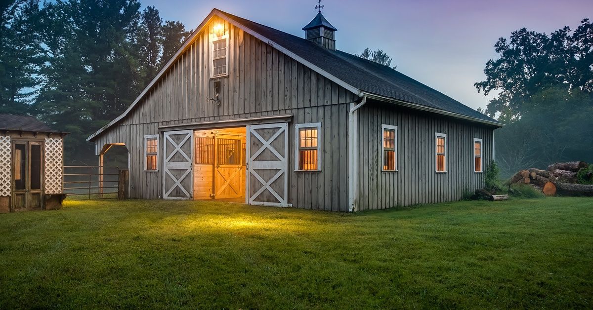 A large gray barn in a lush field at dusk. An exterior light is on outside of the barn, and the wind