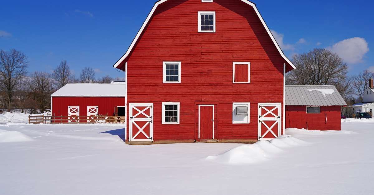 A tall, red, traditional barn with windows and white trim on a farm in the wintertime with snow on the ground.