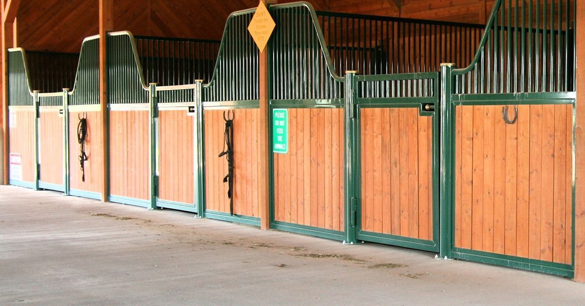 The interior of an open wooden post-frame building, with three horse stables with green trim.