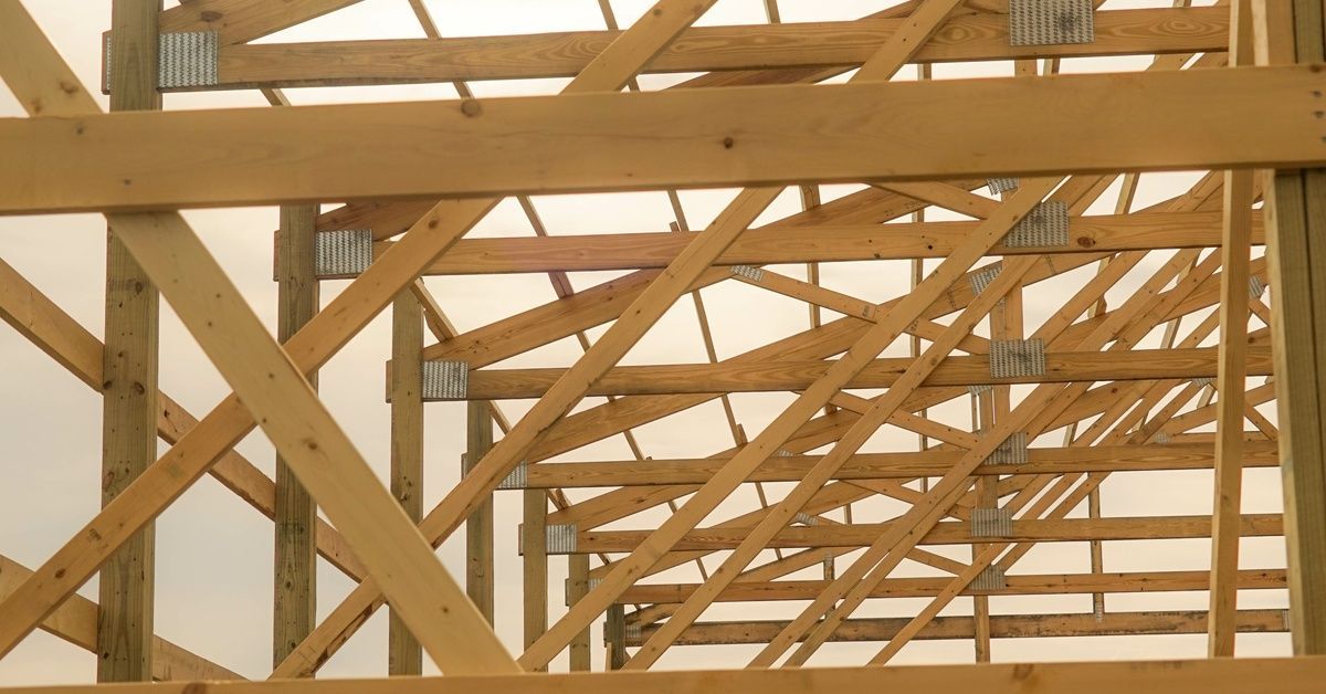 The wooden frame and bones of a pole building sit on a grassy field with the sun shining through the open structure.