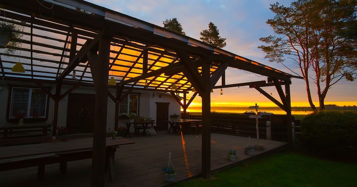 The wood frame of an unfinished patio, connected to a house, with pole construction. The sun sets in the distance.