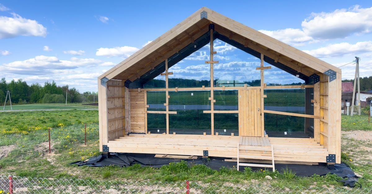 The wooden frame of an unfinished barndominium, sitting on a fenced-in, grassy section of land with a blue sky above.