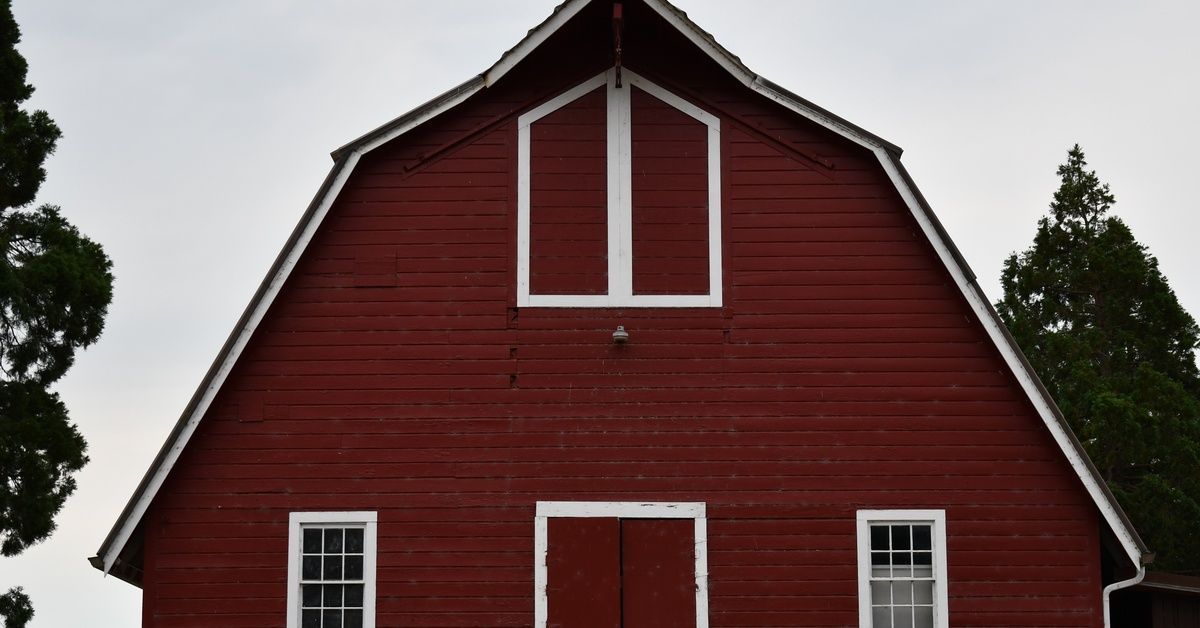 The front of a large barn with red paneling and white trim. The sky is gray, and two trees are behind the barn.