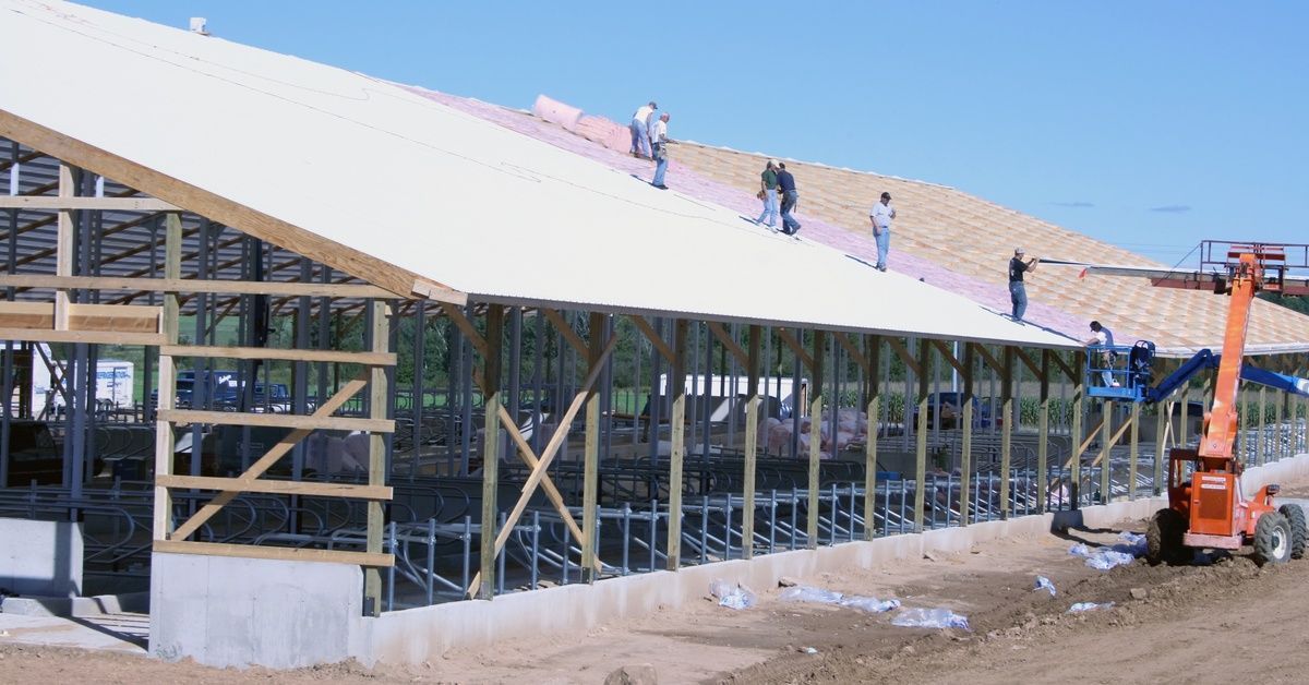 A large wood frame for a barn that is under construction. Multiple builders are working on the roof.