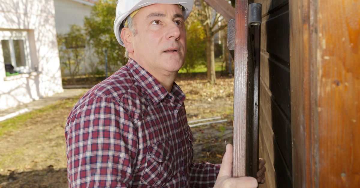 A man wearing a button-up plaid shirt and a white hard hat, lifting a piece of wood against a wood building.