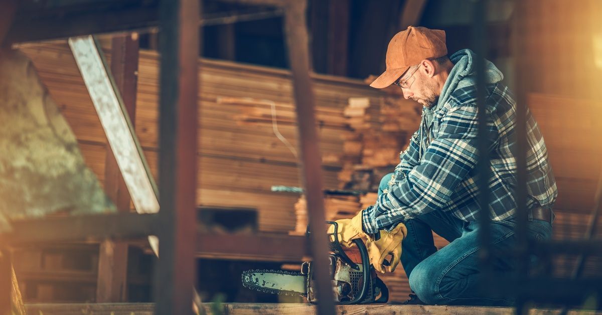 A man wearing industrial gloves and safety glasses, sawing a piece of wood. He is next to a woodworking station.