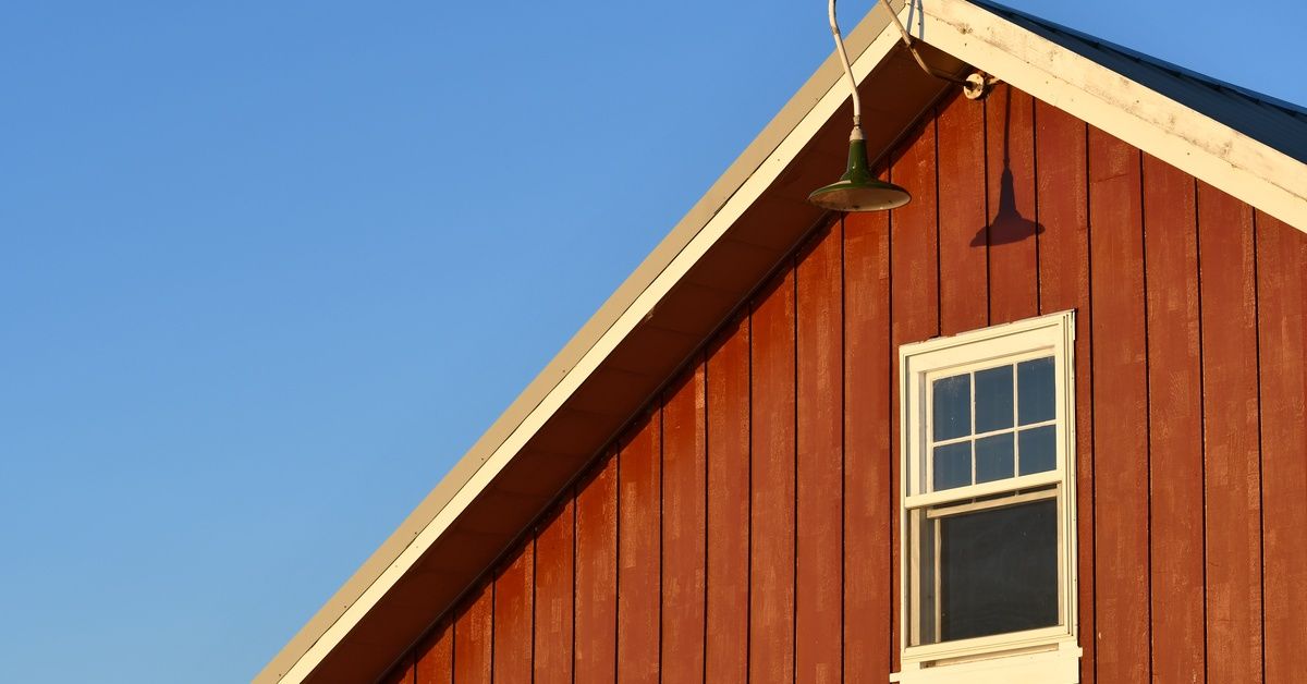A close-up of the top of a red barn. A light fixture extends from the roof and hangs above a single window.