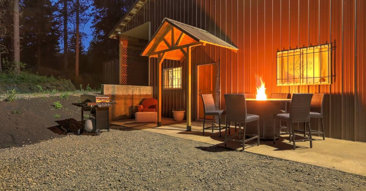 The exterior of a large metal barn at night. A round fire pit and outdoor furniture sit in front of the barn.