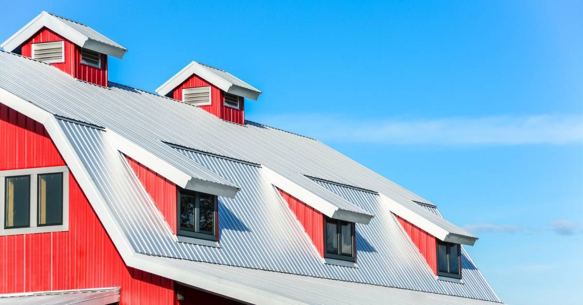 The angular metal roof of a red barn with several small windows. The sky behind the barn is clear and blue.