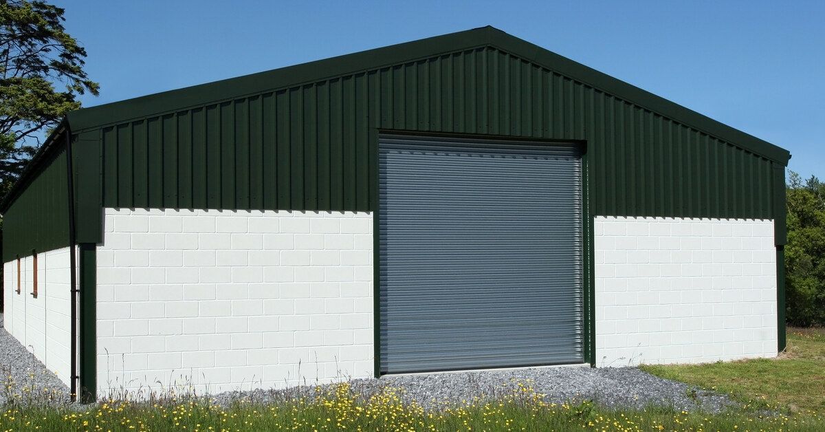 A barn with a white brick exterior and a green metal roof. There is a large gray garage door in the center of one side.