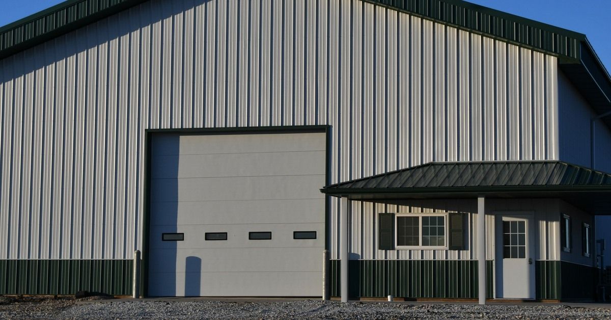 The exterior of a large gray and green metal building with a peaked roof. The building has a garage door in the front.