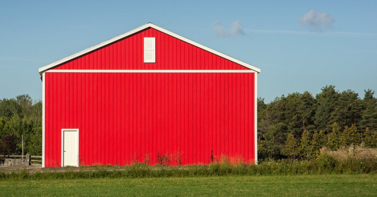 The exterior of a bright red metal barn. The barn has a white door near one wall and a small window by the roof.