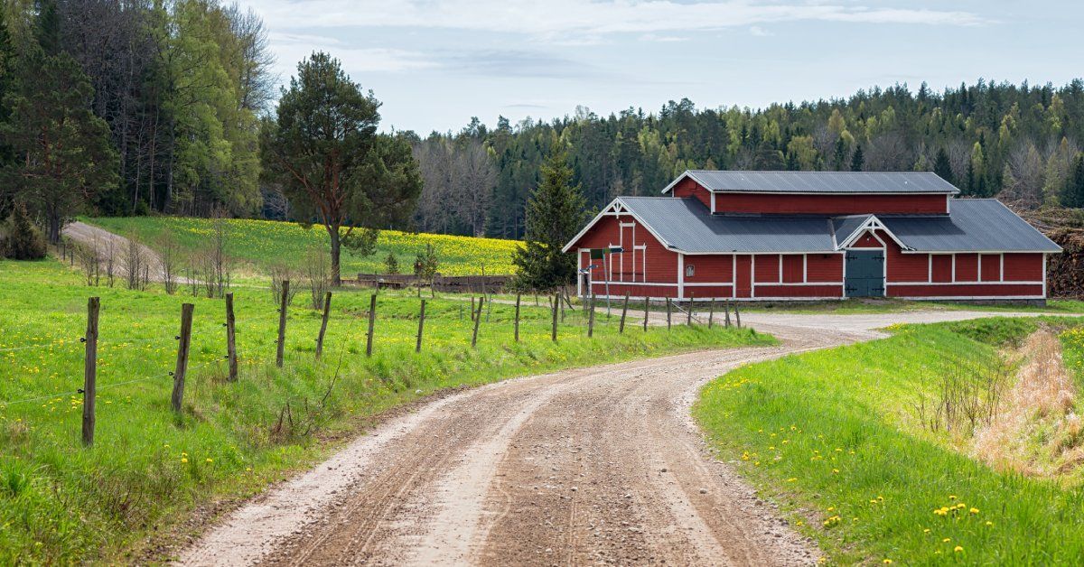 A large red and white barn surrounded by mature trees and open fields. A dirt road leads to the barn.