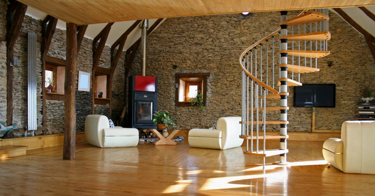 The interior of a rustic room with wood floors, a spiral staircase, and stone walls. There is a fireplace in the corner.