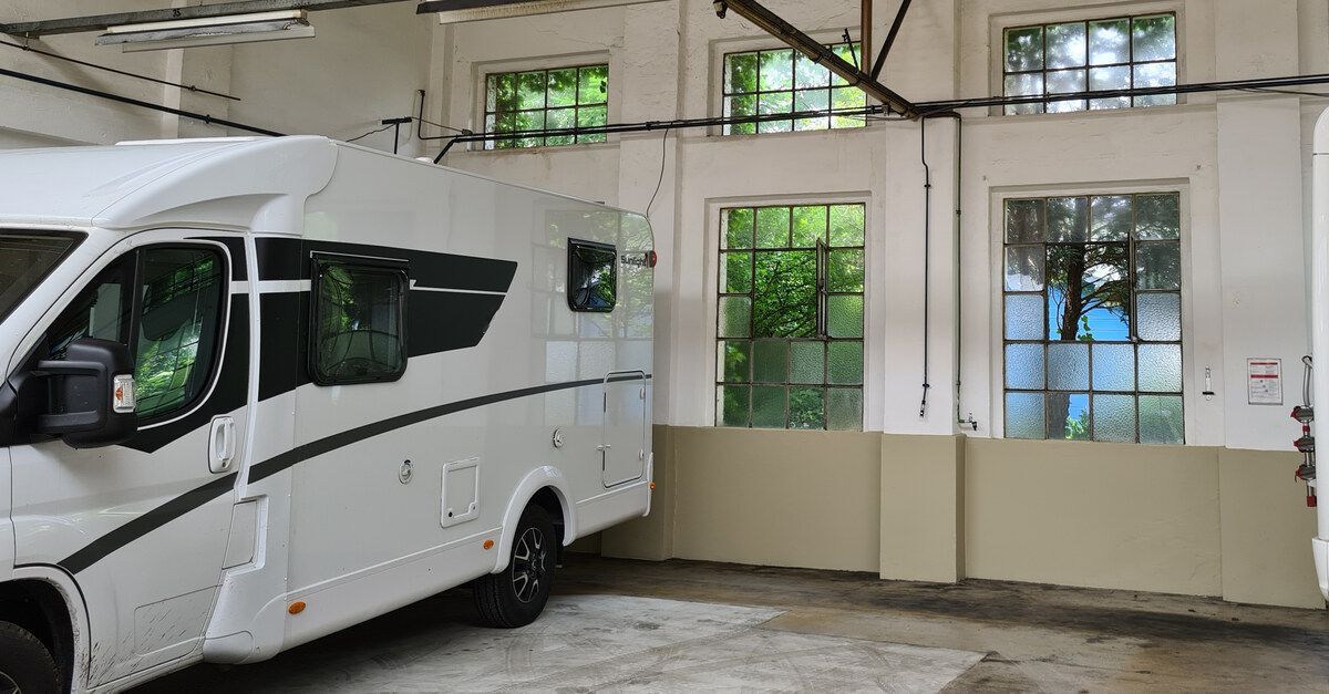 A black and white RV parked inside a large garage. The garage has lights on the ceiling and several windows on one wall.