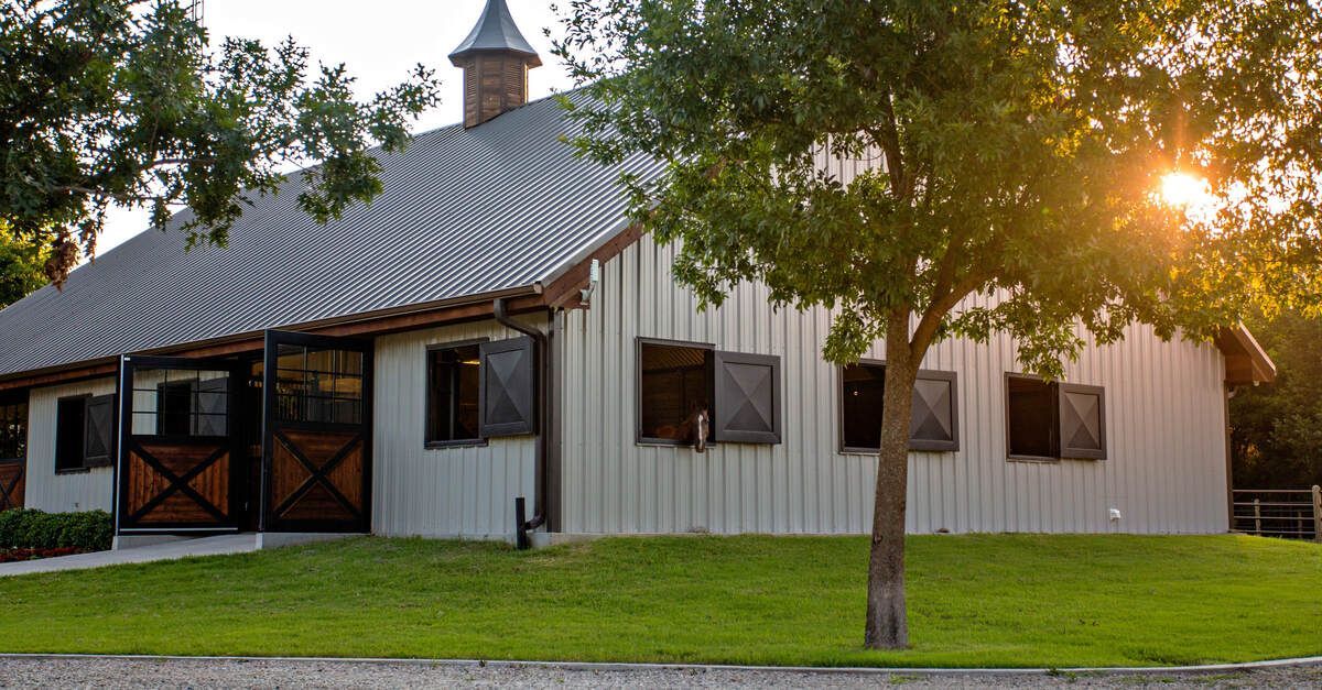 The exterior of a white metal barn with multiple windows. There is a turret on the roof and a horse 