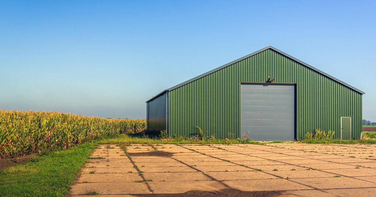 A green metal building with a peaked roof and a large gray garage door surrounded by grass and tall crops.