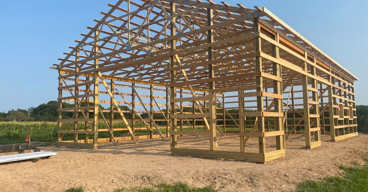The wooden framing of a barn under construction. The structure is sitting on dirt and has a peaked r