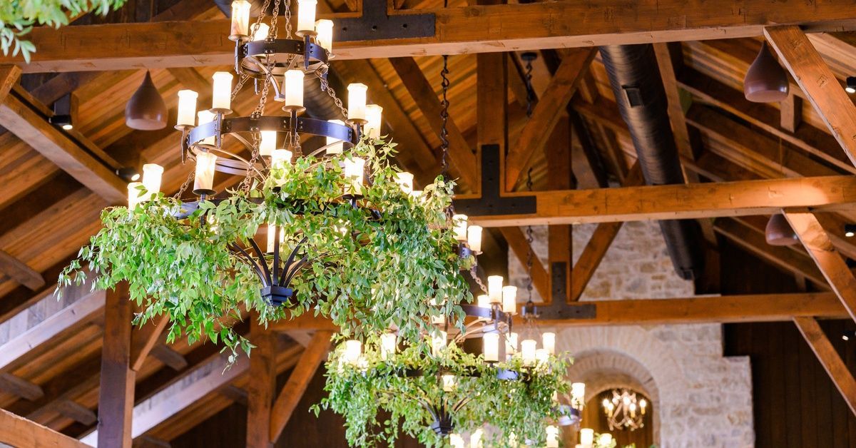 Three multi-tier chandeliers decorated with greenery and suspended from the wooden beams of a vaulted ceiling.