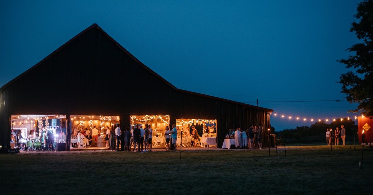 A barn set up for an evening party and filled with people. The barn has a peaked roof and twinkling 