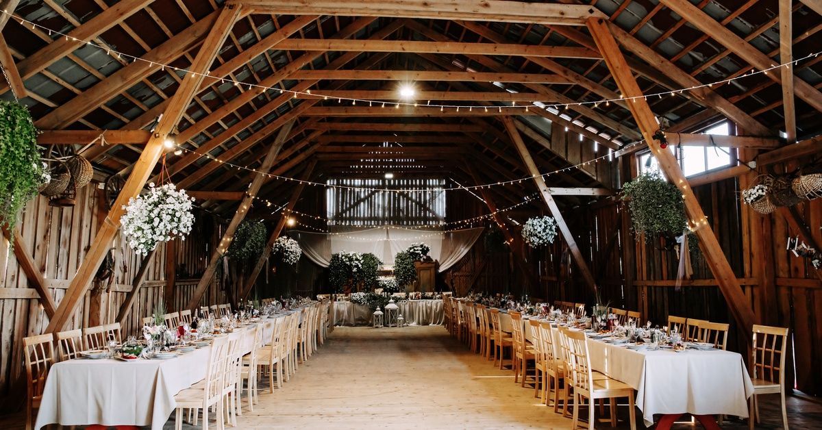 The interior of a barn with three long tables covered with white tablecloths and flowers and lights suspended from wooden beams.
