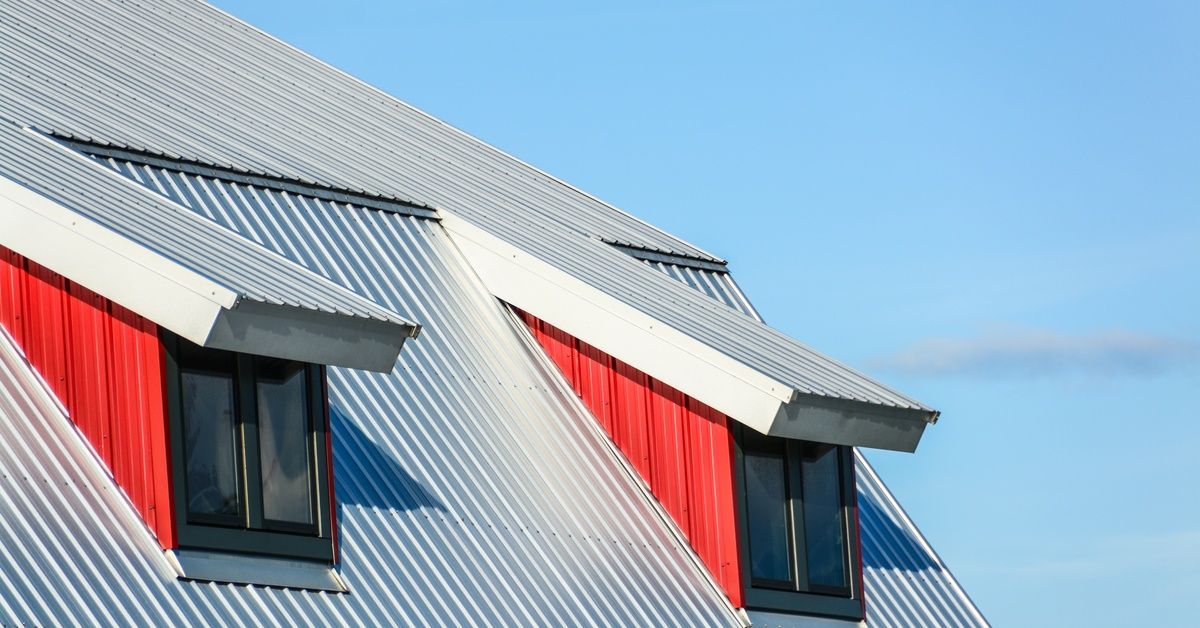 A close-up of a barn's metal roof with two black and red dormer windows with a blue sky in the background.