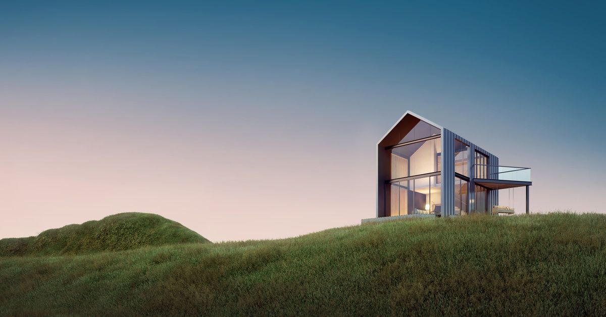 A modernized barn-style house with one side made of glass and with a large second-floor deck sits on a grassy hill.