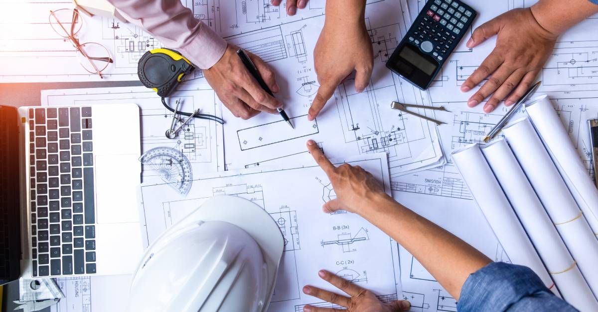 A close up shot of various building blueprints and three construction workers pointing and taking notes on them.