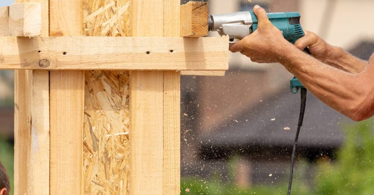 A close-up of a person wearing a hat holding a power drill to drill into a wooden post of a new building going up.