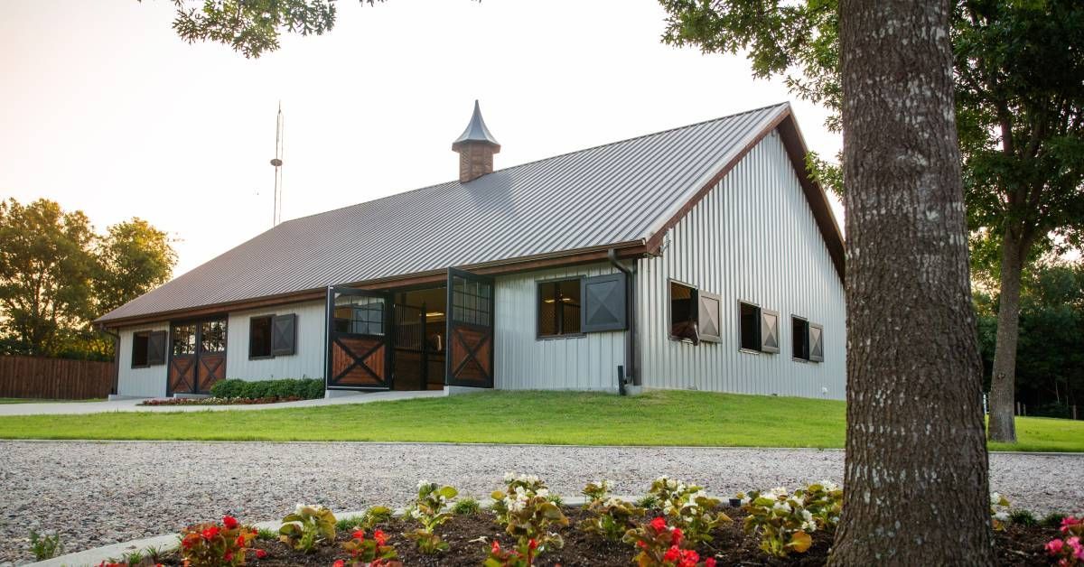 A beautiful pole barn building outfitted with wide double doors and windows. A horse sticks its head out a window.