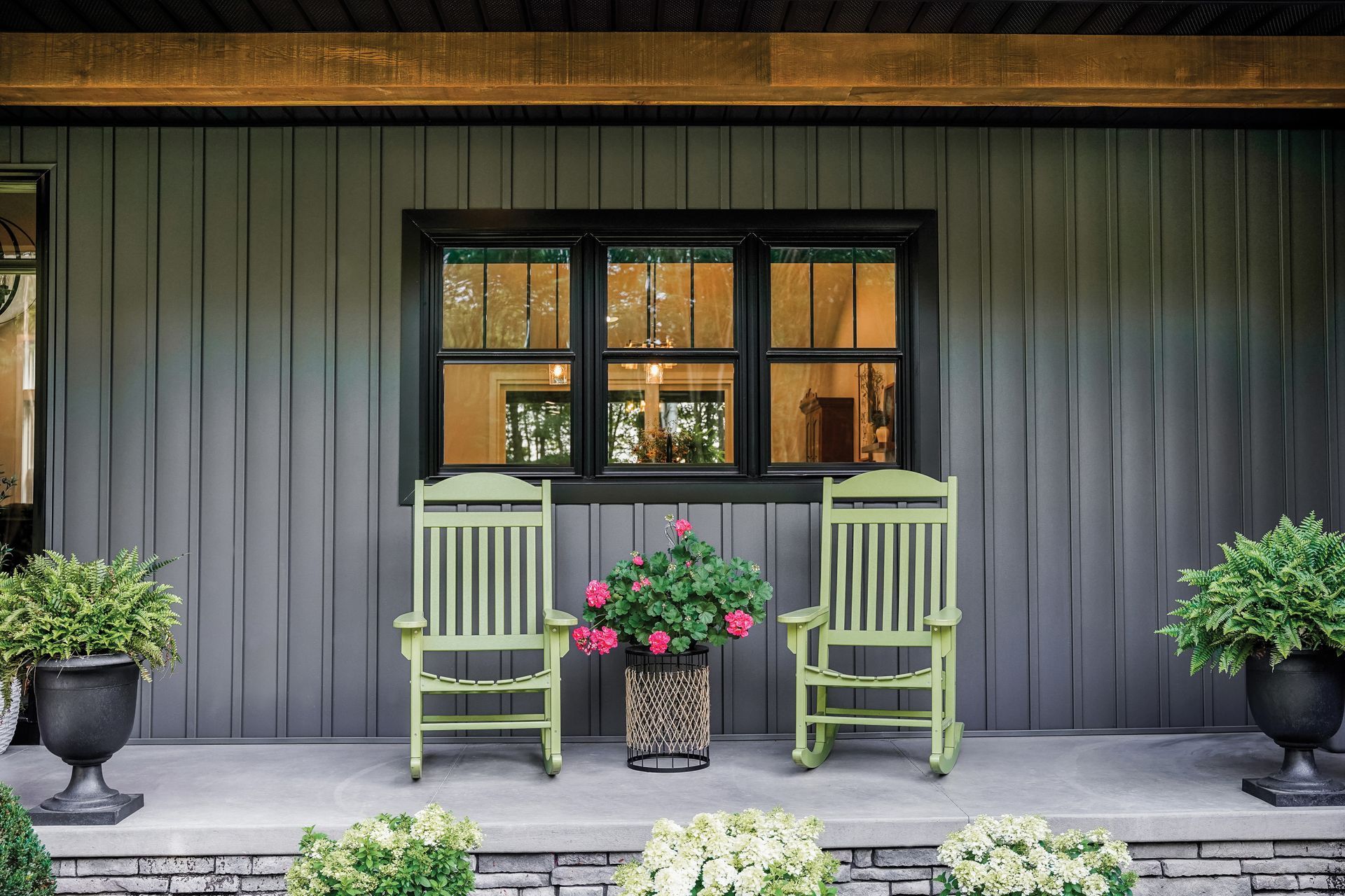 Two green rocking chairs sit on a porch, flanking a potted plant, under a dark window against gray siding.