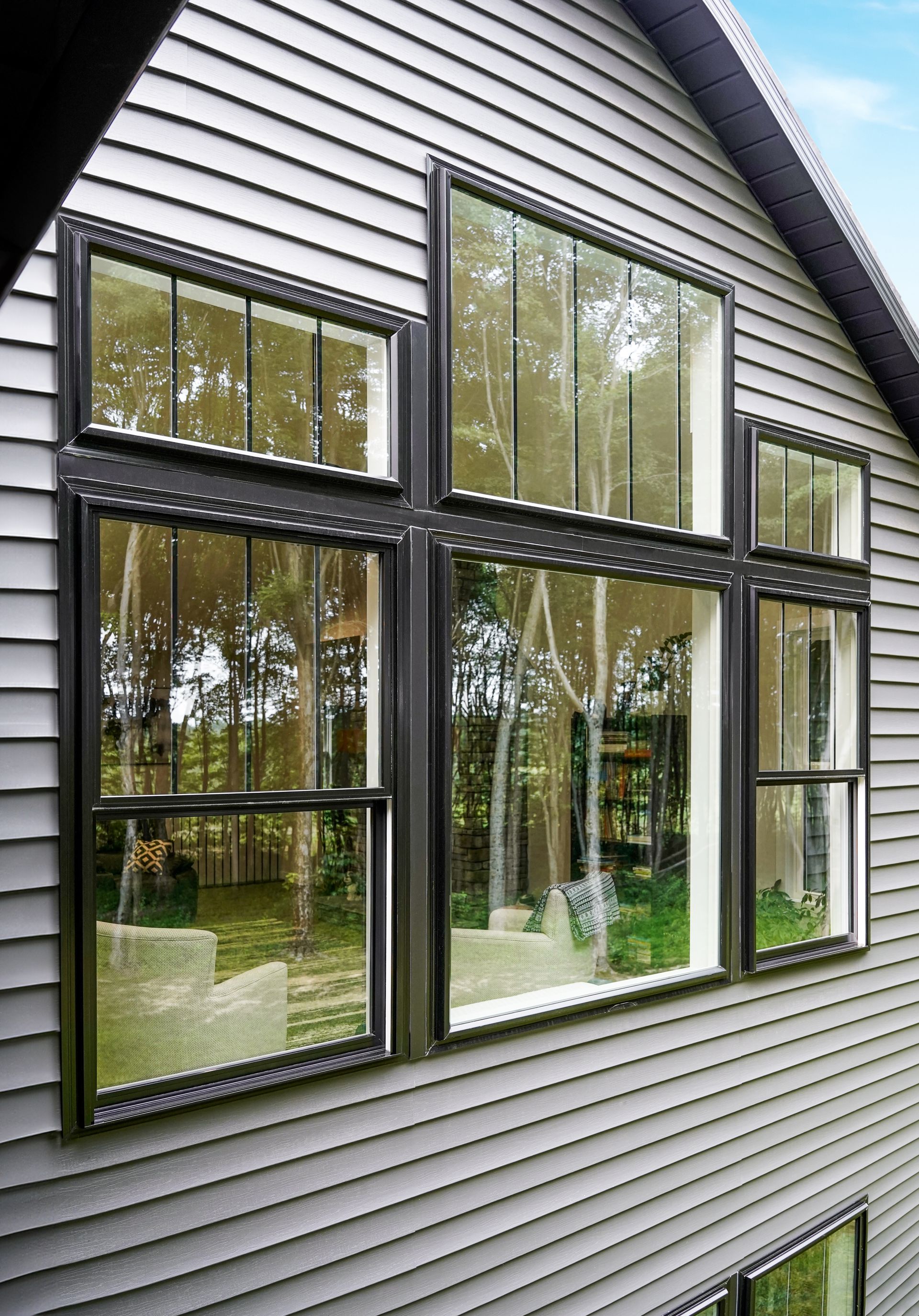 Gray house exterior with dark-framed windows reflecting trees. Vertical siding and angled roofline are also visible.