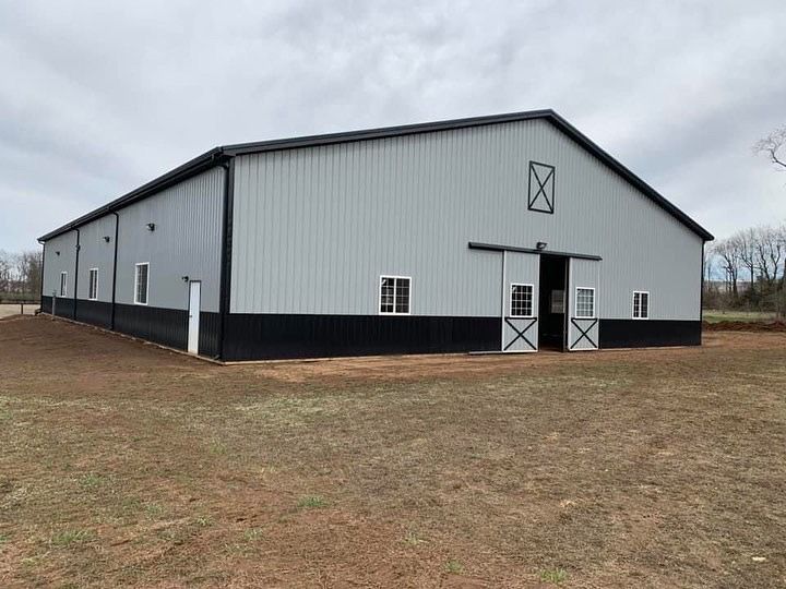 Large metal barn, silver walls, black lower section, black trim, sliding door, white windows, set in a grassy field under a cloudy sky.