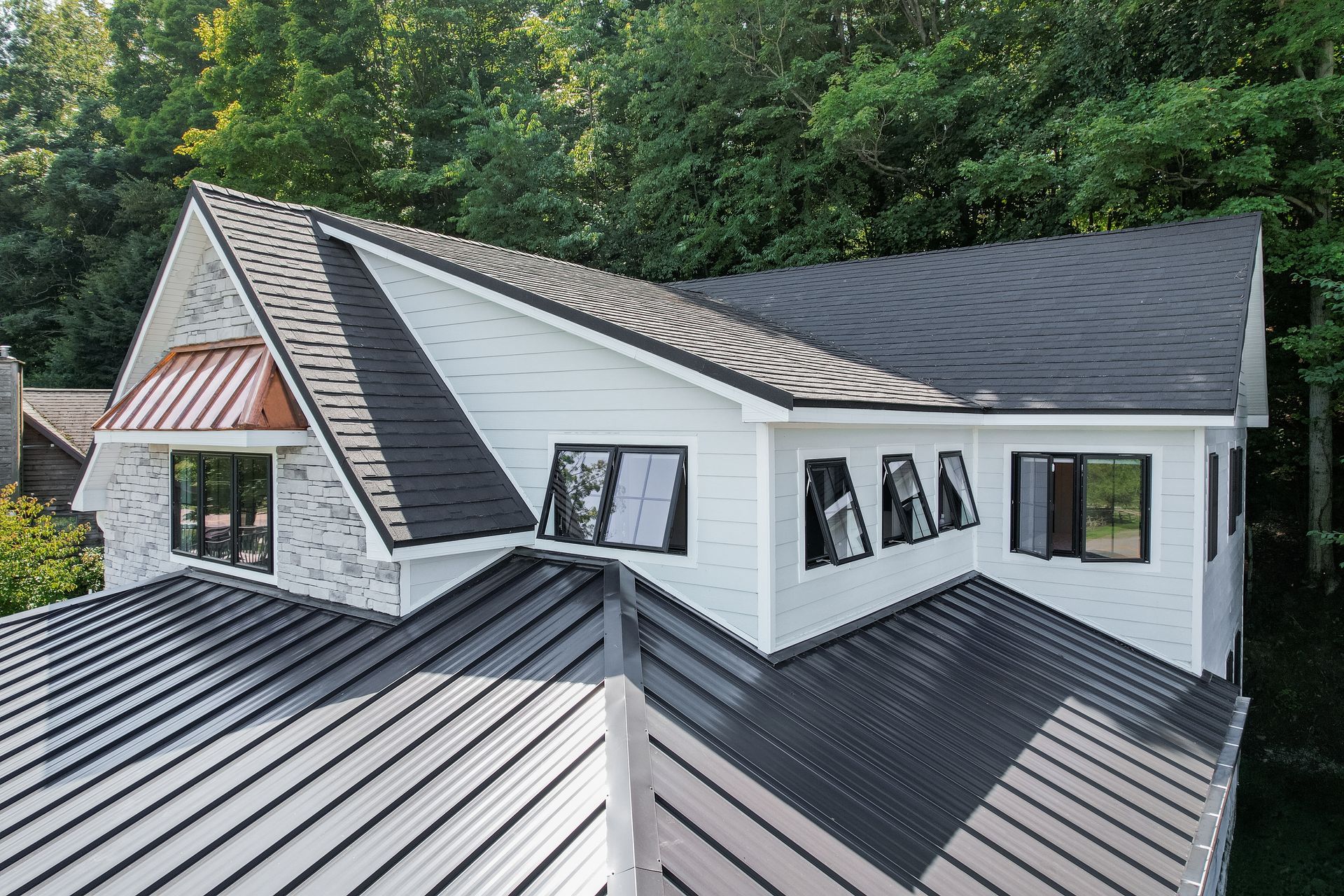White house with dark metal roof, black-framed windows, and copper accent. Set against a backdrop of green trees.
