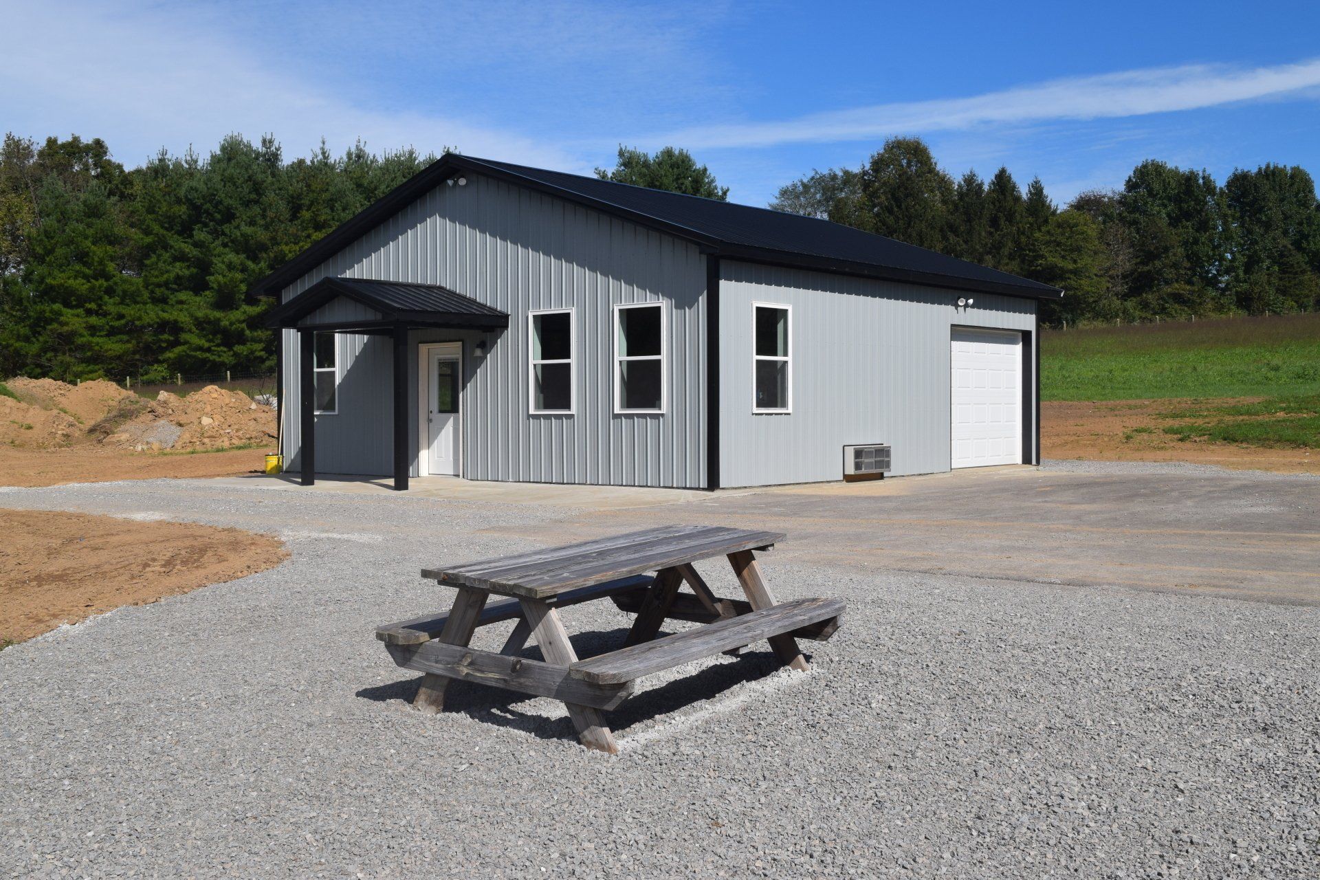 A gray metal building with a black roof, a picnic table, and a gravel driveway under a blue sky.