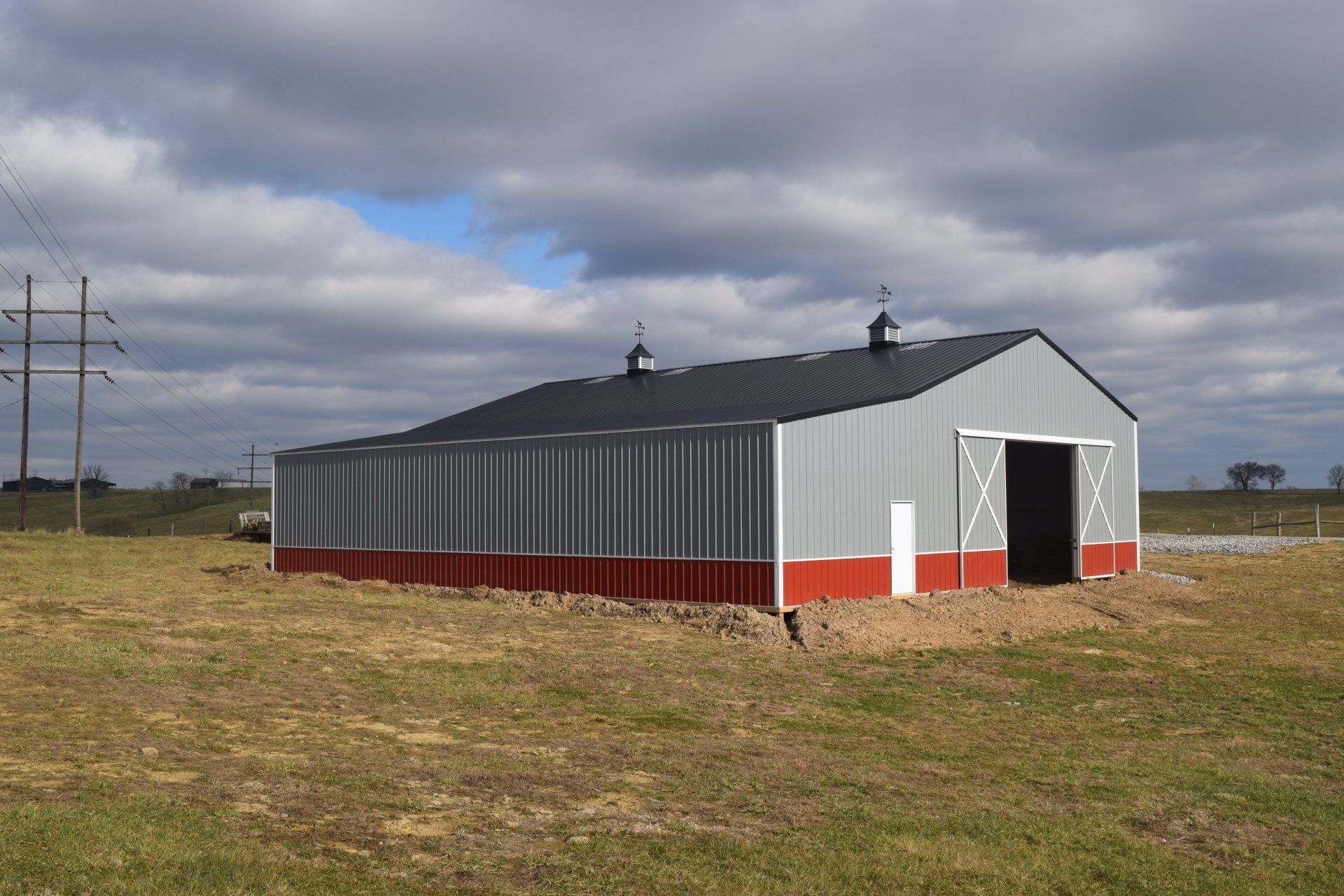 A large metal barn with a gray body and red base sits in a field under a cloudy sky. A dark roof and open door are visible.
