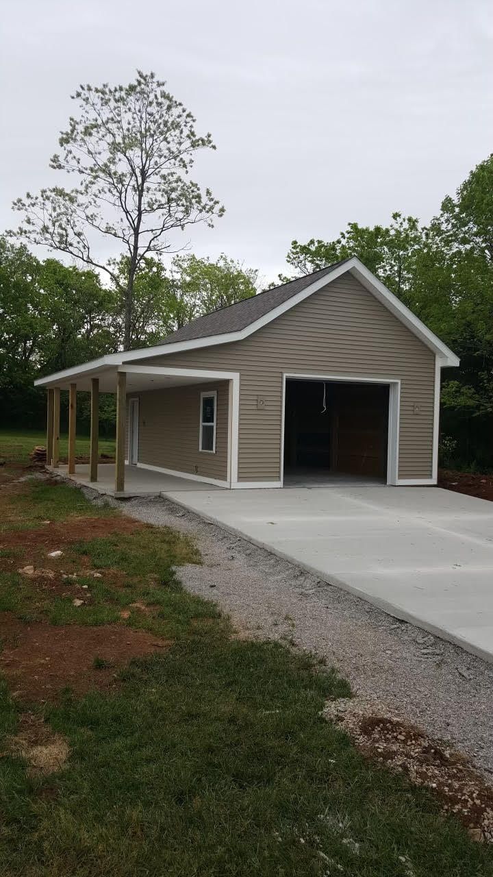 Tan and white garage with carport, concrete driveway, and surrounding trees.