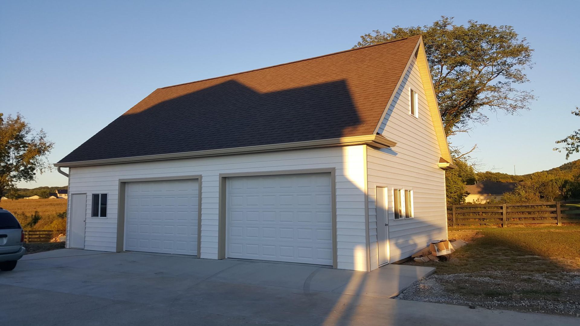 Two-car garage with white siding and brown roof, set against a rural backdrop under a clear sky.