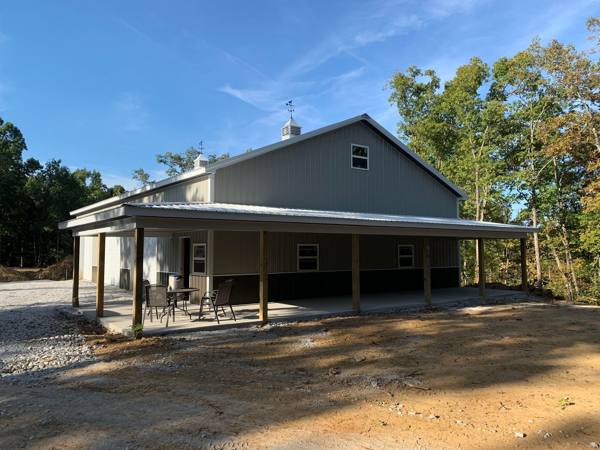 A gray barn with a porch and seating area in a rural setting on a sunny day.