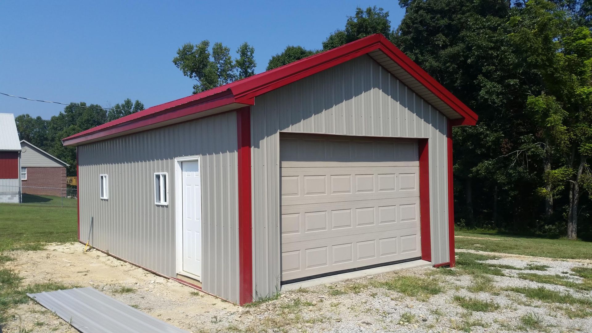 Tan metal garage with a red roof and trim, featuring a garage door and a white door, set on a grassy area with trees.