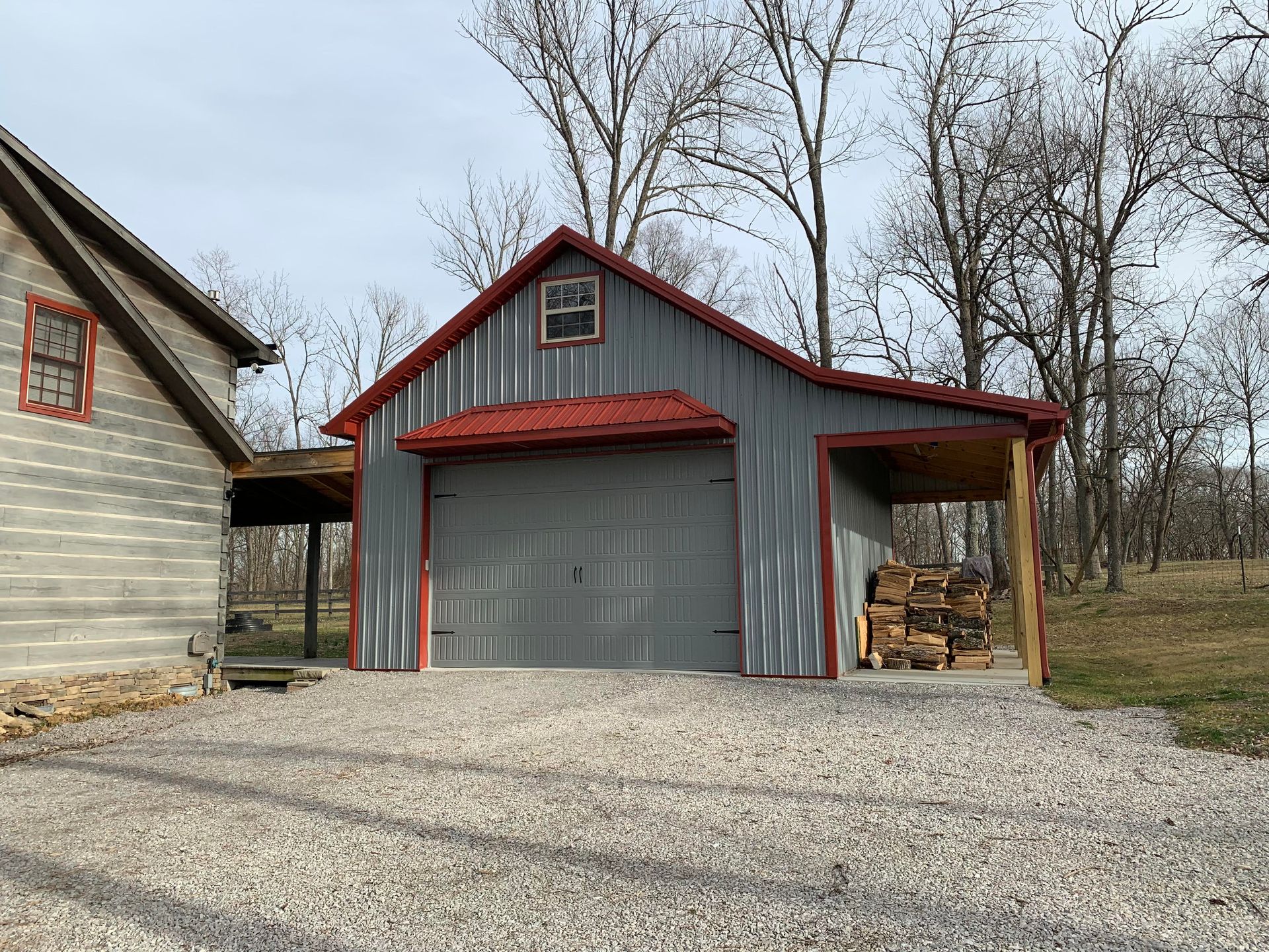 Gray garage with red trim and roof, attached to a covered wood storage area. Building is on a gravel driveway, next to a light-colored house.