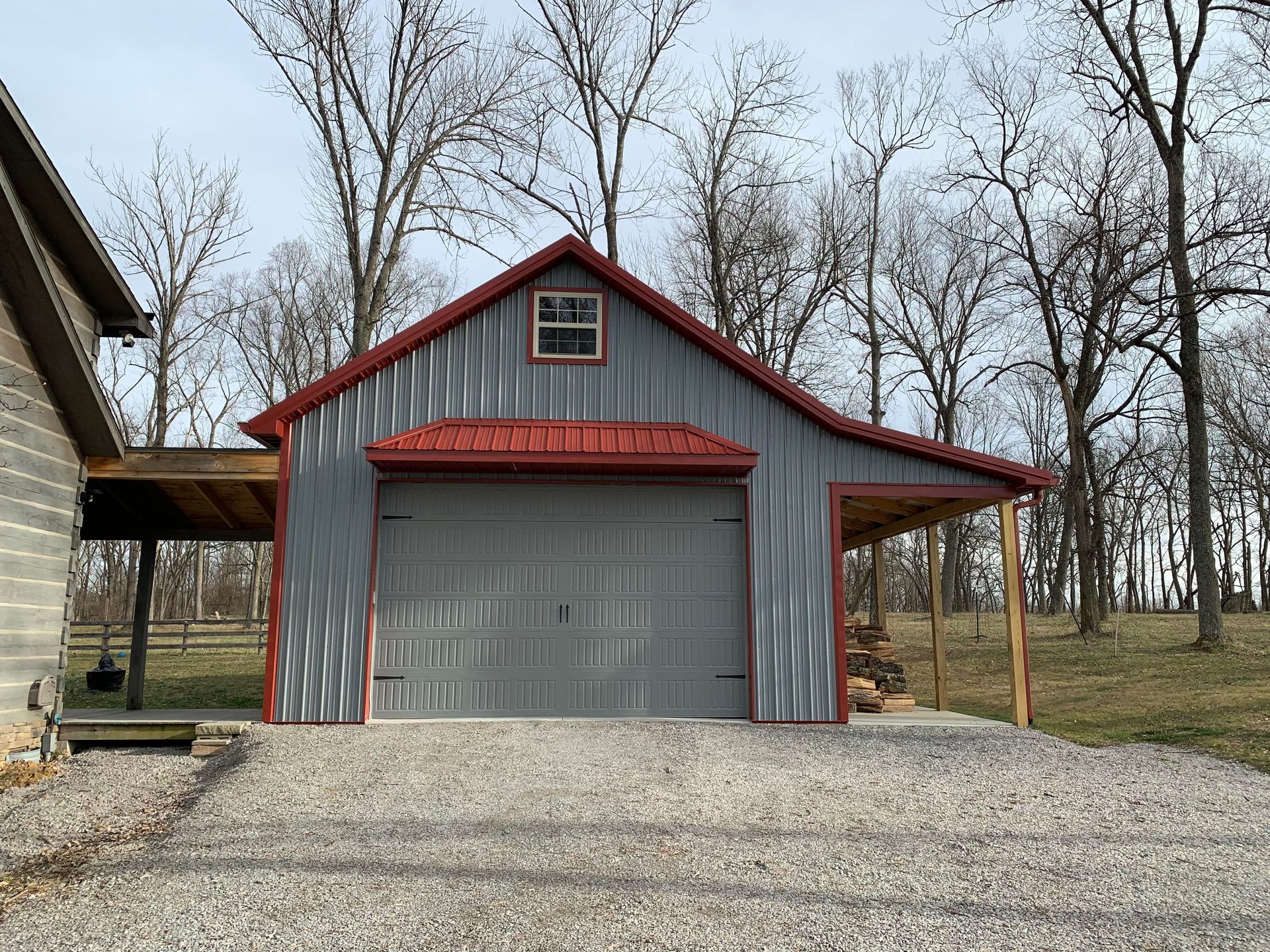 Gray metal garage with red trim and roof, small window, and attached wooden carport, set against trees.