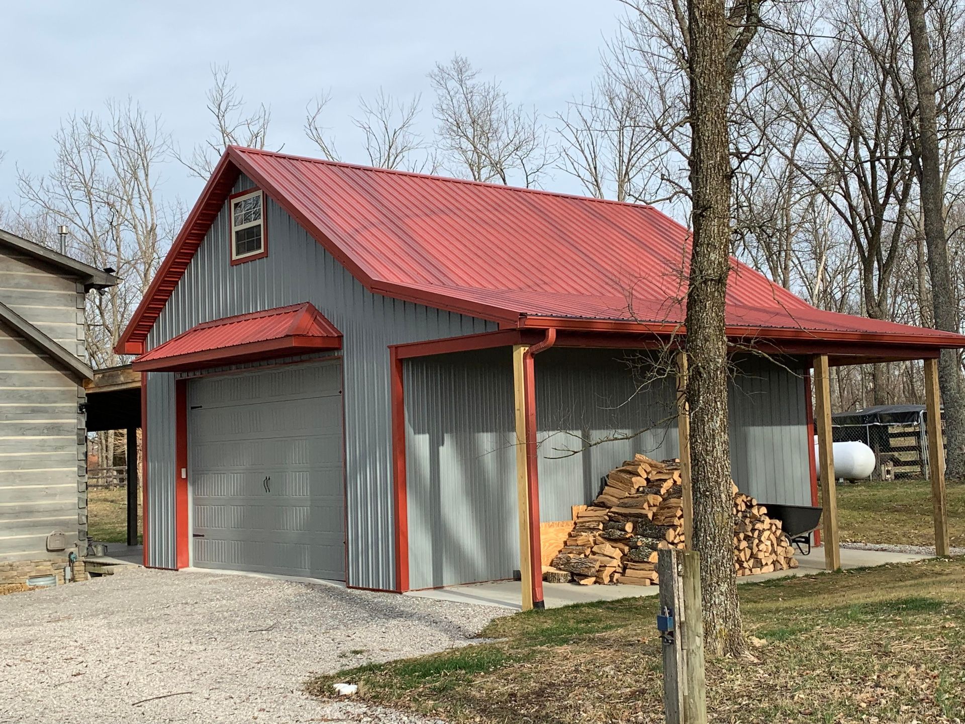 A gray metal garage with a red roof and trim. There's firewood stacked to the right and a small window in the gable.