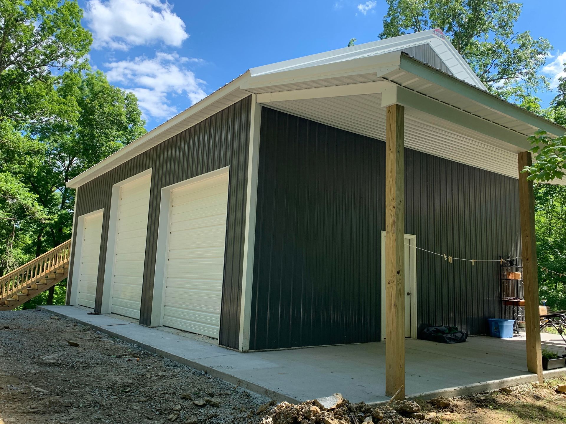 A dark gray metal garage with three white garage doors, a porch, and a wooden staircase, set in a wooded area.