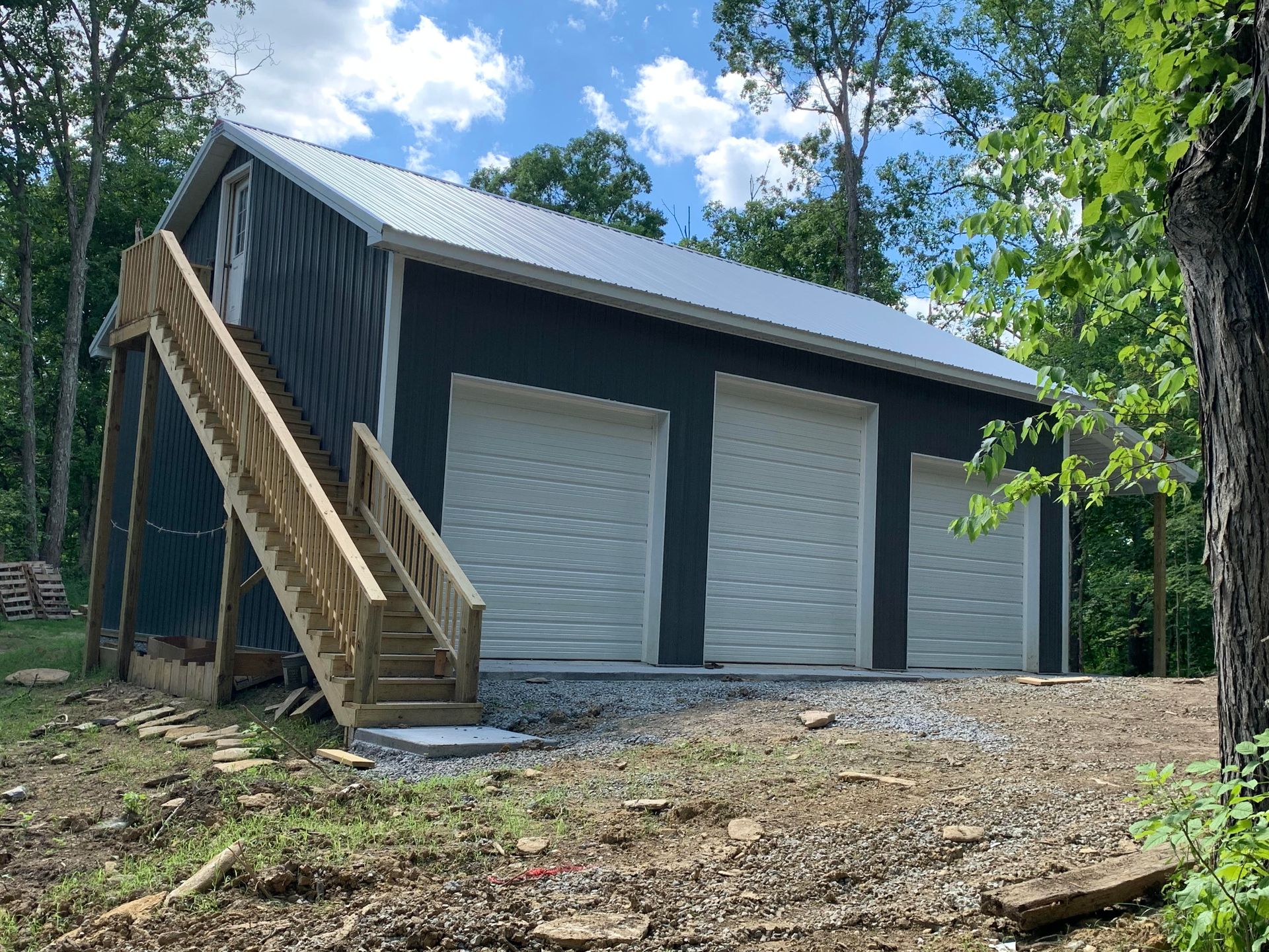 Dark blue three-bay garage with a white metal roof and a wooden staircase leading to an upper level. The building is surrounded by trees.