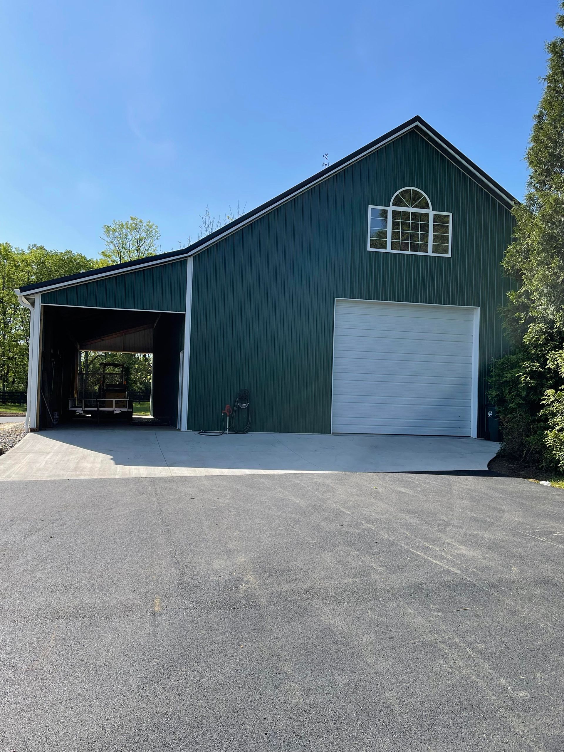 Green barn with white garage door and open carport. Dark gravel driveway in foreground, blue sky background.